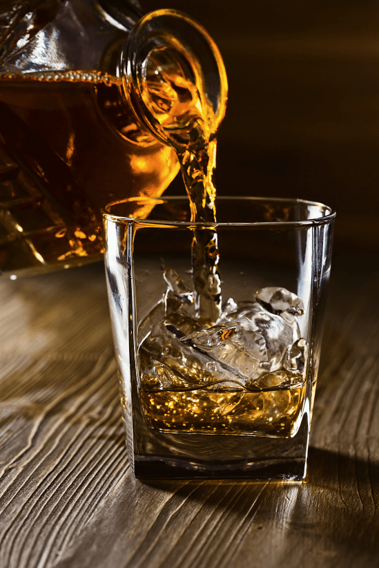 A glass filled with ice as whiskey is poured from a decanter, captured in warm lighting on a wooden surface.